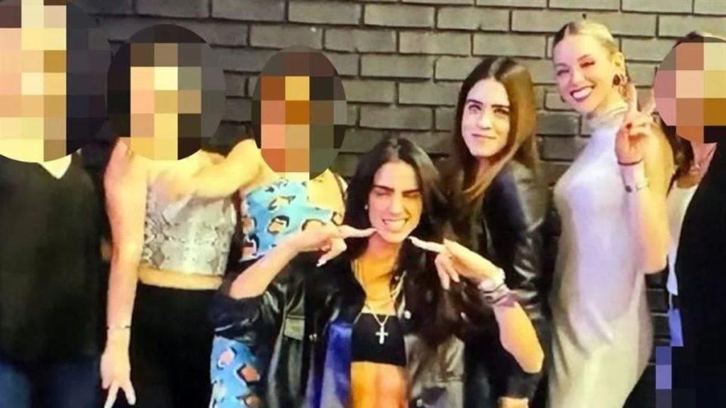 Group of Young Friends Posing for a Photo in Front of a Brick Wall; Center Figure Makes a Playful Finger Gesture Near Her Lips While Others Smile. | Ciudadano Seguros