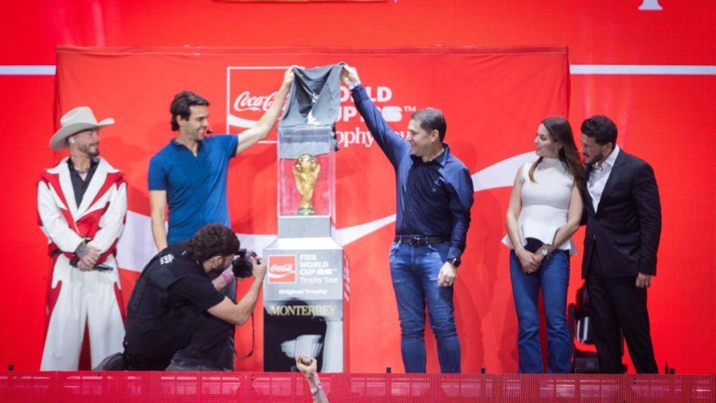 Six People Stand on a Red Stage, Lifting a Covered Trophy As a Photographer Crouches to Capture the Moment on a Coca-cola Backdrop. | Ciudadano Seguros