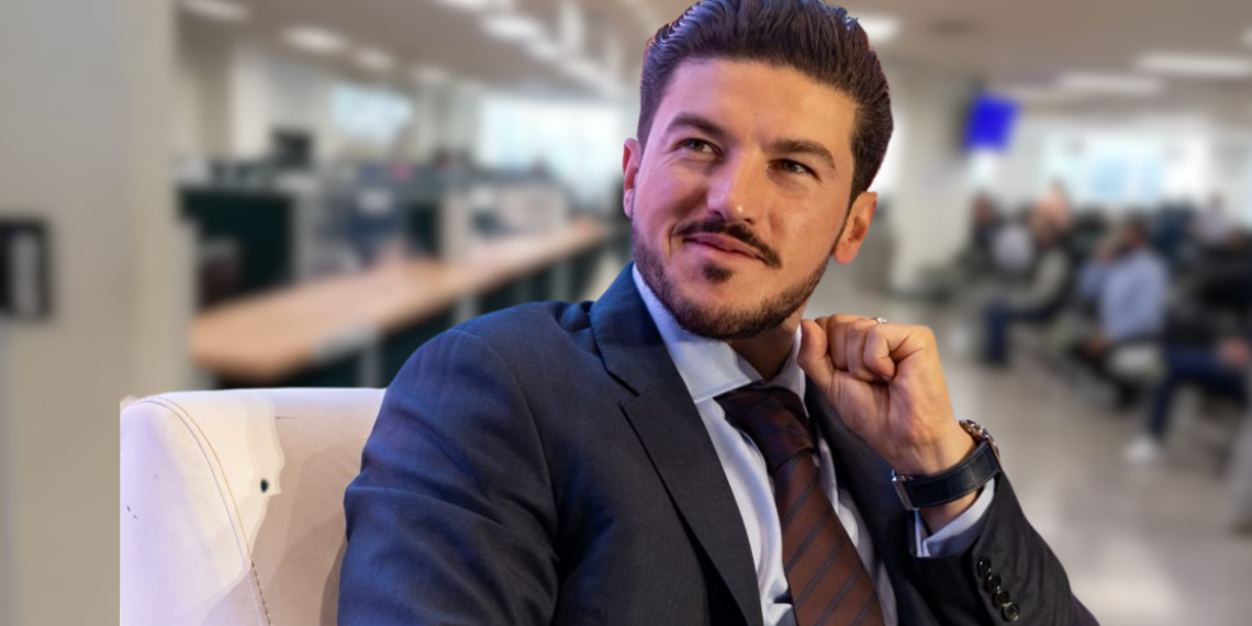 Man in a Navy Suit and Brown Tie Sits on a White Chair in a Bright, Modern Office Lobby, Looking Thoughtful. | Ciudadano Seguros