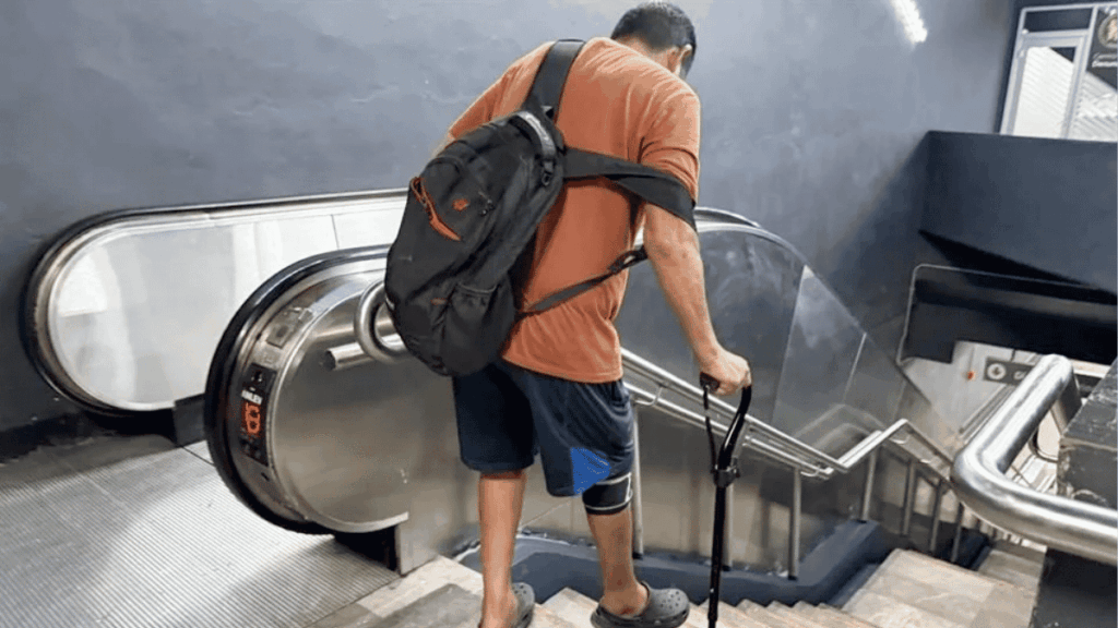 Fallas En El Metro | Ciudadano Seguros Man in an Orange Shirt and Blue Shorts with a Backpack Stands on an Escalator, Using a Cane for Support. | Ciudadano Seguros