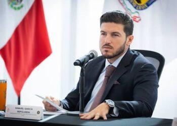 Man in a Dark Suit and Tie Sits at a Press Conference Table, Speaking into a Microphone with Flags in the Background. | Ciudadano Seguros