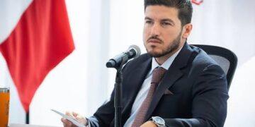Man in a Dark Suit and Tie Sits at a Press Conference Table, Speaking into a Microphone with Flags in the Background. | Ciudadano Seguros