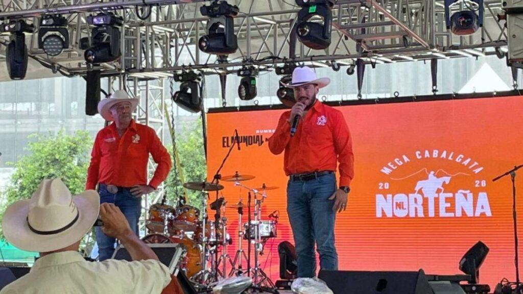 Two Men in Red Shirts and White Cowboy Hats on a Stage, One Speaking into a Microphone, with Drums Behind Them and an Orange Norteña Banner in the Background; Foreground Audience Member Wears a Beige Hat. | Ciudadano Seguros