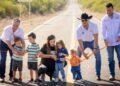 Group of Adults and Children Participate in a Ribbon-cutting on a Rural Road Opening Ceremony with an Orange Ribbon Stretched Across the Road Waist-high. | Ciudadano Seguros
