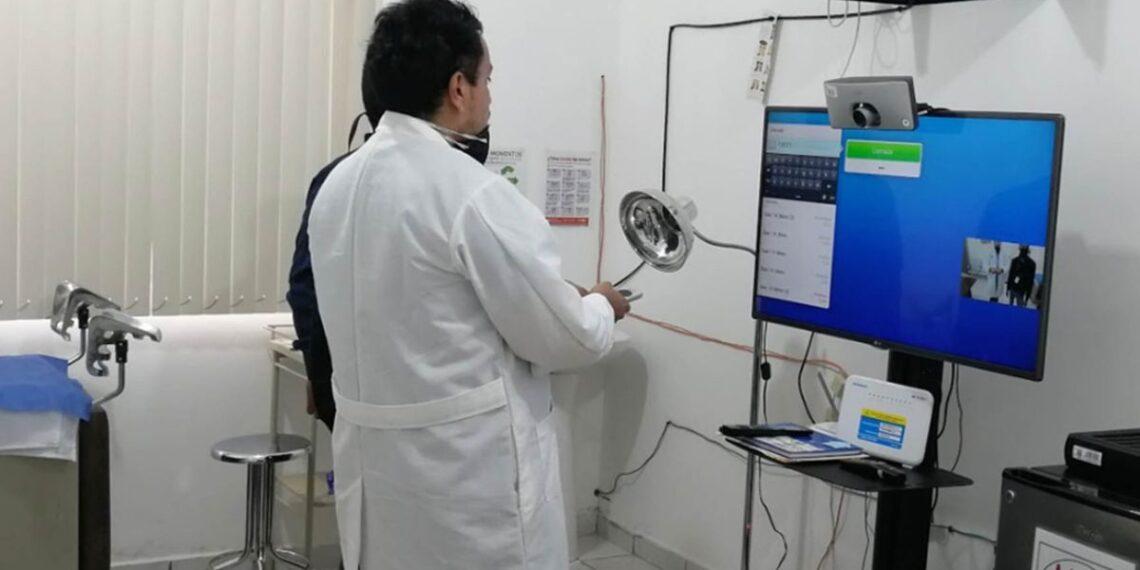 Doctor in a White Coat Stands by a Computer Setup in a Clinic, Holding a Magnifying Lamp During a Telemedicine Session with a Monitor and Webcam Nearby. | Ciudadano Seguros