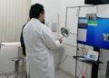 Doctor in a White Coat Stands by a Computer Setup in a Clinic, Holding a Magnifying Lamp During a Telemedicine Session with a Monitor and Webcam Nearby. | Ciudadano Seguros