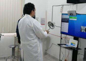 Hospitales-de-nuevo-leon | Ciudadano Seguros Doctor in a White Coat Stands by a Computer Setup in a Clinic, Holding a Magnifying Lamp During a Telemedicine Session with a Monitor and Webcam Nearby. | Ciudadano Seguros